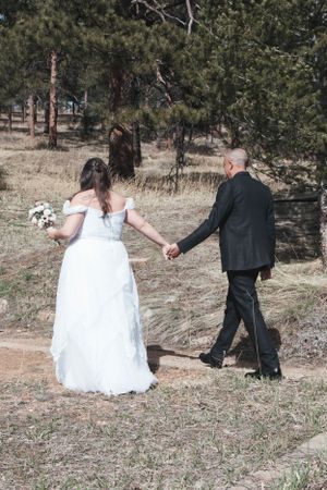 A couple walking hand in hand on a wooded path, with the bride wearing a flowing white dress.