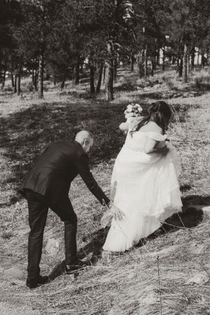 A black and white photo shows a couple navigating down a wooded hillside in wedding attire.