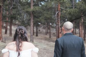 A couple in wedding attire viewed from behind as they walk through a pine forest.