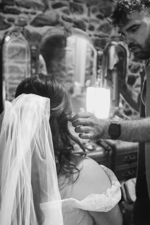 A hairstylist works on styling a bride's hair while she wears her wedding veil in a black and white photo.