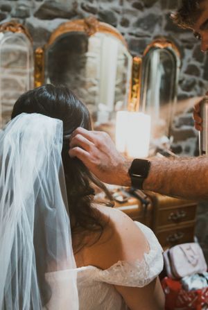 A bride having her veil adjusted while getting ready in a stone-walled room with ornate gold mirrors.