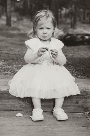A small child in a white dress and shoes sits on wooden planks in a black and white photograph.