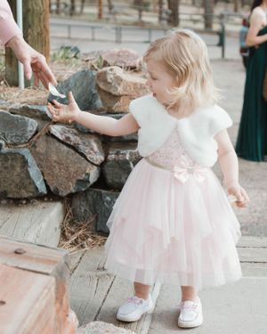 A small child in a pink tulle dress and white bolero reaches towards a stone wall while wearing white shoes.