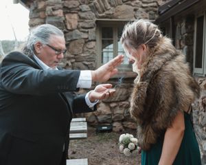 A touching moment during a wedding ceremony as an officiant gestures emotionally to someone in a fur wrap outdoors.