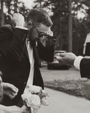 Black and white photo showing a person in sunglasses and suit jacket leaning forward with white roses at an outdoor event.