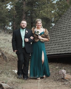 A couple in formal attire walking along a mountain path near a rustic cabin.