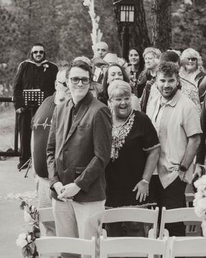 A black and white photograph showing a group of people standing by white folding chairs at an outdoor gathering.