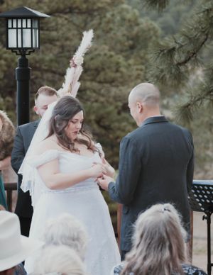 An outdoor wedding ceremony shows a couple exchanging rings under a lantern and pine trees.