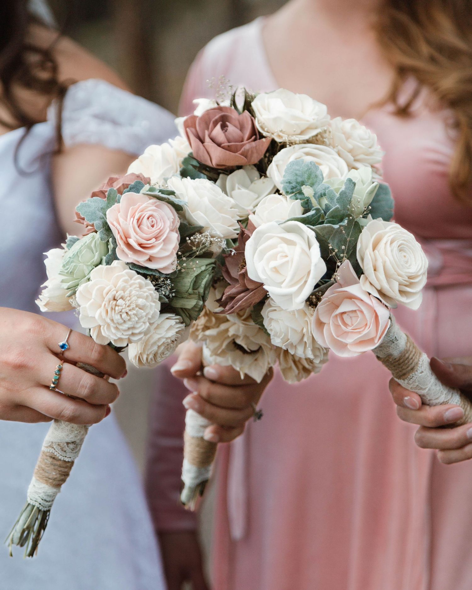 Wedding bouquets with blush pink and white roses wrapped in twine are held by hands in pink and white dresses.