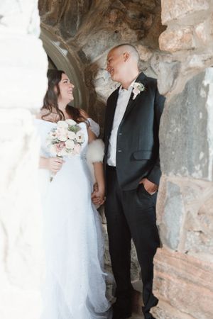 A bride in a white gown and groom in a black tuxedo share a romantic moment in a rustic stone archway.