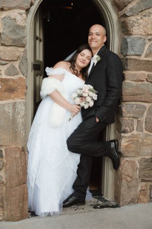 A bride and groom pose together in a stone church doorway during their wedding day.
