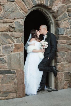 A romantic wedding couple shares a kiss in a stone archway while the bride holds a delicate bouquet of flowers.