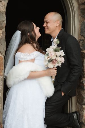 A joyful wedding couple laughs together in a stone doorway while the bride holds a pink and white bouquet.