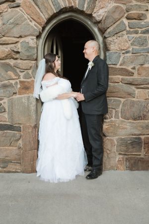 A couple stands in a stone archway during their wedding, with the bride in white and groom in black formal attire.