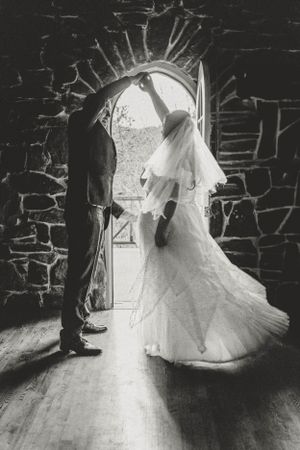 A black and white wedding photo showing a couple in an arched stone doorway with dramatic lighting and flowing dress.