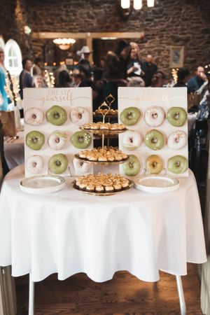 Wedding dessert table with tiered display stand and donuts arranged on a white tablecloth in a rustic venue.
