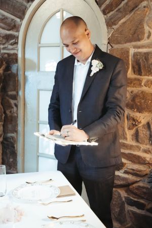 A groom in a black suit signs documents at a stone-walled venue near an arched doorway.