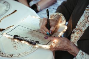 Hands signing an ornate wedding document illuminated by string lights.