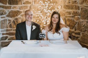 Newlyweds sitting at a signing table with a stone wall and twinkle lights behind them.