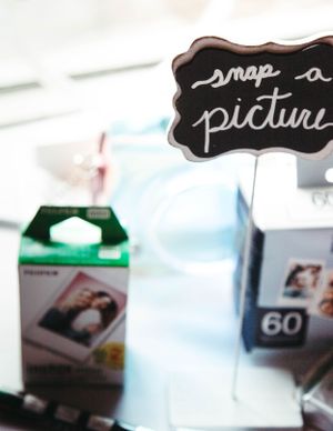A chalkboard sign reading 'snap a picture' with Polaroid film boxes blurred in the background.