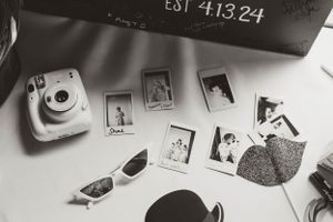 Black and white photo showing instant polaroid photos scattered on a surface with sunglasses and an Instax camera.