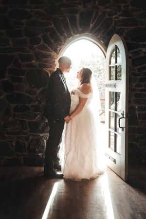 A couple stands in silhouette against bright light streaming through an arched stone church doorway on their wedding day.