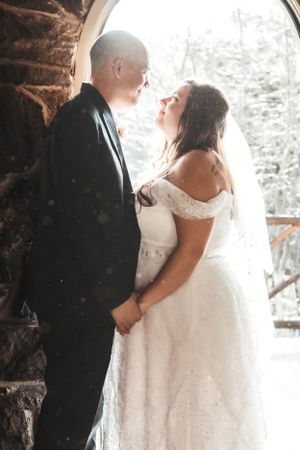 A romantic wedding photo of a couple in formal attire standing close together against a rustic stone wall background.