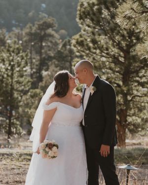 A couple shares a romantic moment in their wedding attire among tall pine trees on a sunny mountain day.