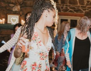 Person with long braids wearing a floral dress dances and holds a drink at a dimly lit party venue.
