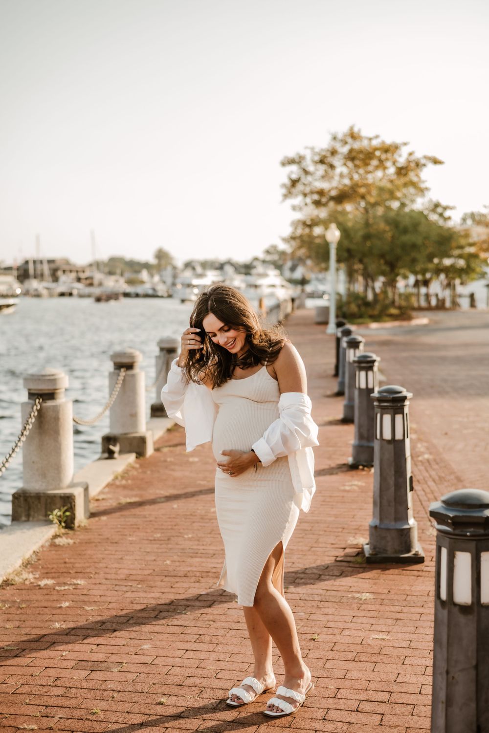 Pregnant woman in white dress walking along waterfront path during sunset.