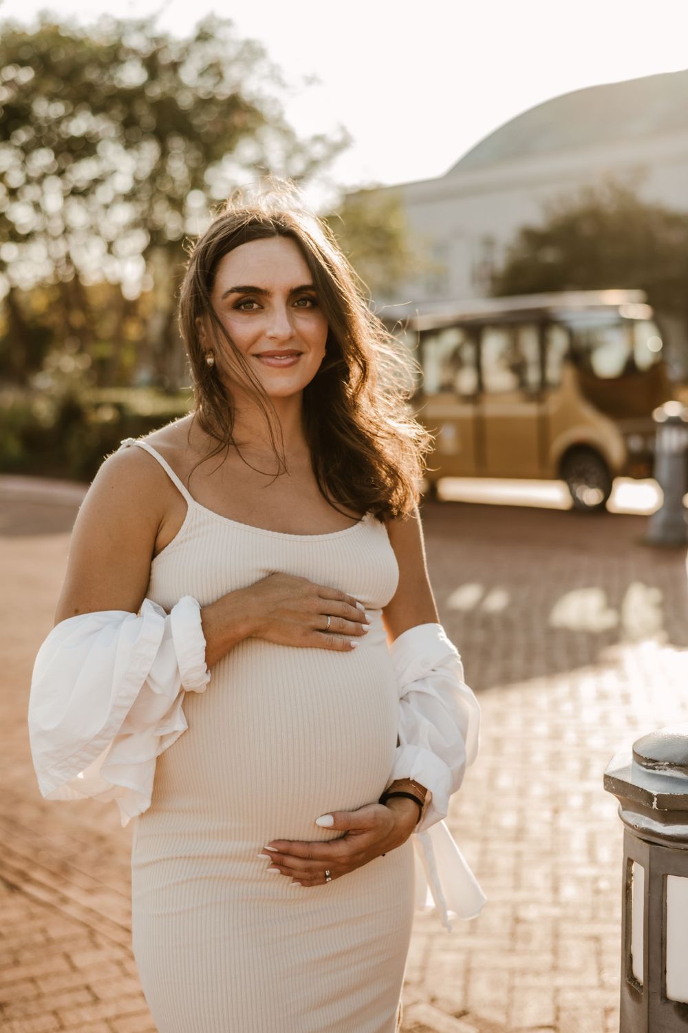 Expectant mother in white dress poses during golden hour photoshoot near vintage bus.