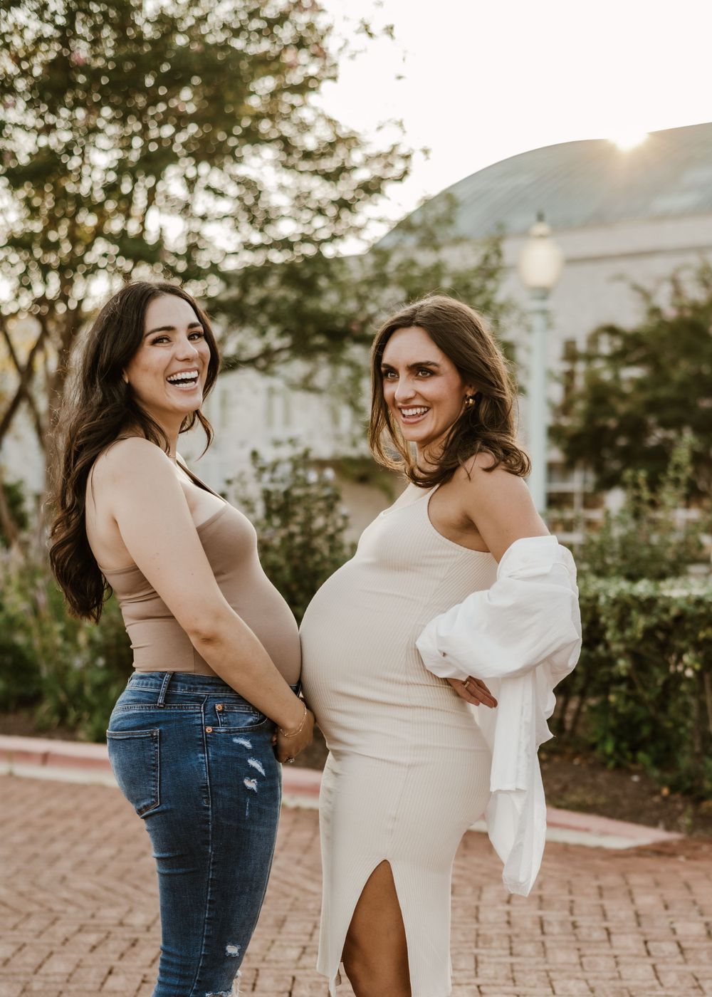 Two women showing off baby bumps while smiling during sunset maternity photoshoot.