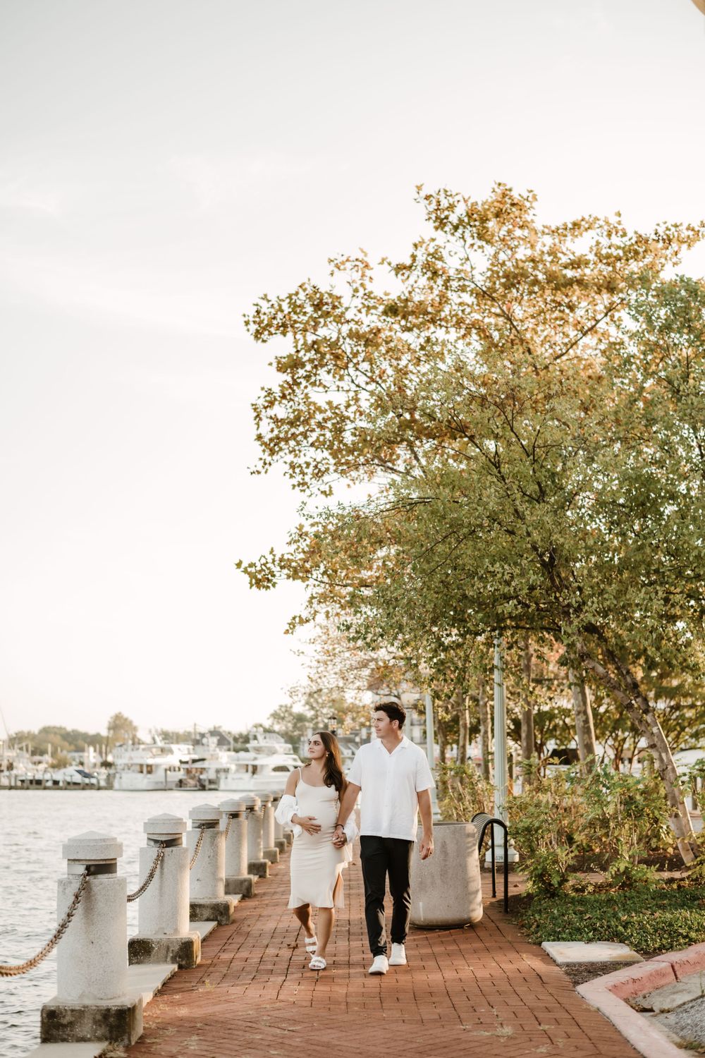 Expecting couple walking along waterfront path with boats and autumn trees in background.