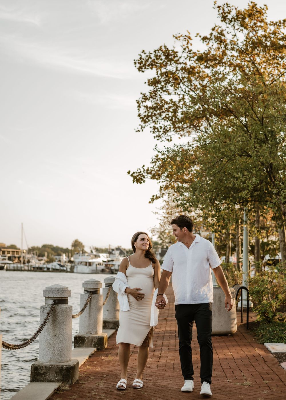 Couple walking along waterfront brick path during sunset maternity photoshoot.