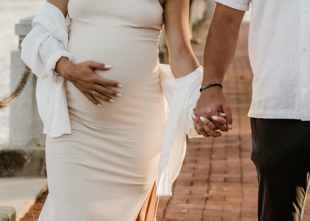 Close-up of hands cradling pregnant belly in white dress during outdoor photoshoot.