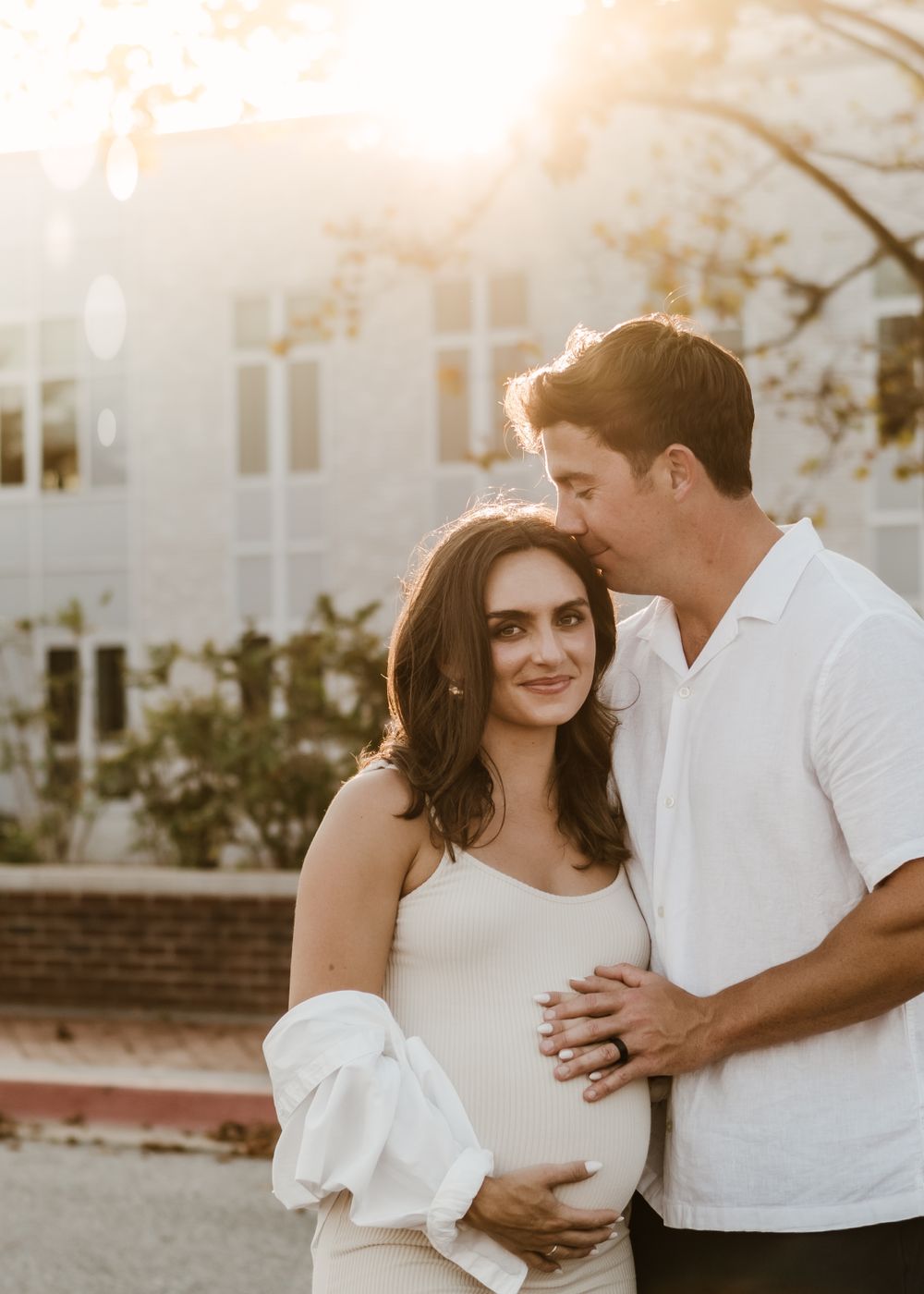 Romantic maternity photoshoot at sunset with couple embracing in front of white building.
