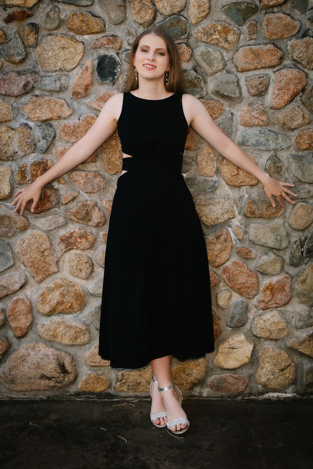 A model in a sleeveless black midi dress poses against a rustic stone wall.
