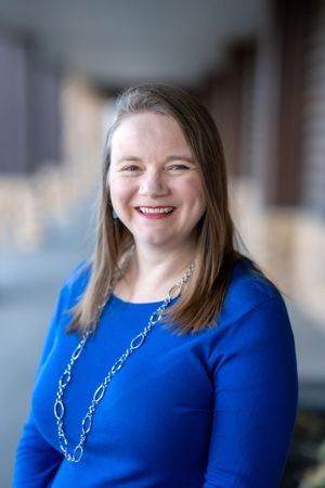 Professional portrait of woman in bright blue top with silver chain necklace against blurred background.