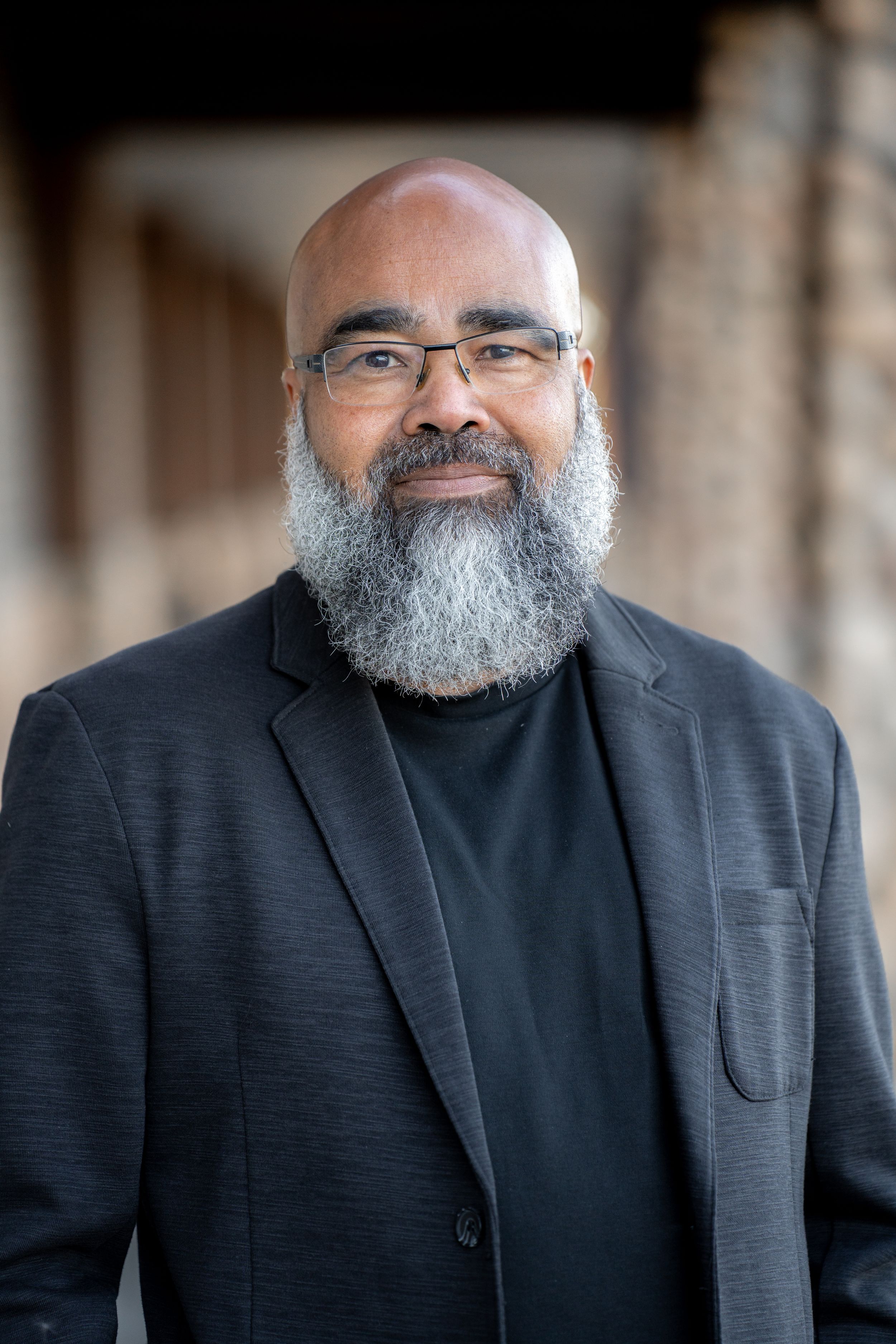 Professional authors portrait of man with gray beard wearing dark suit jacket and glasses against blurred background.