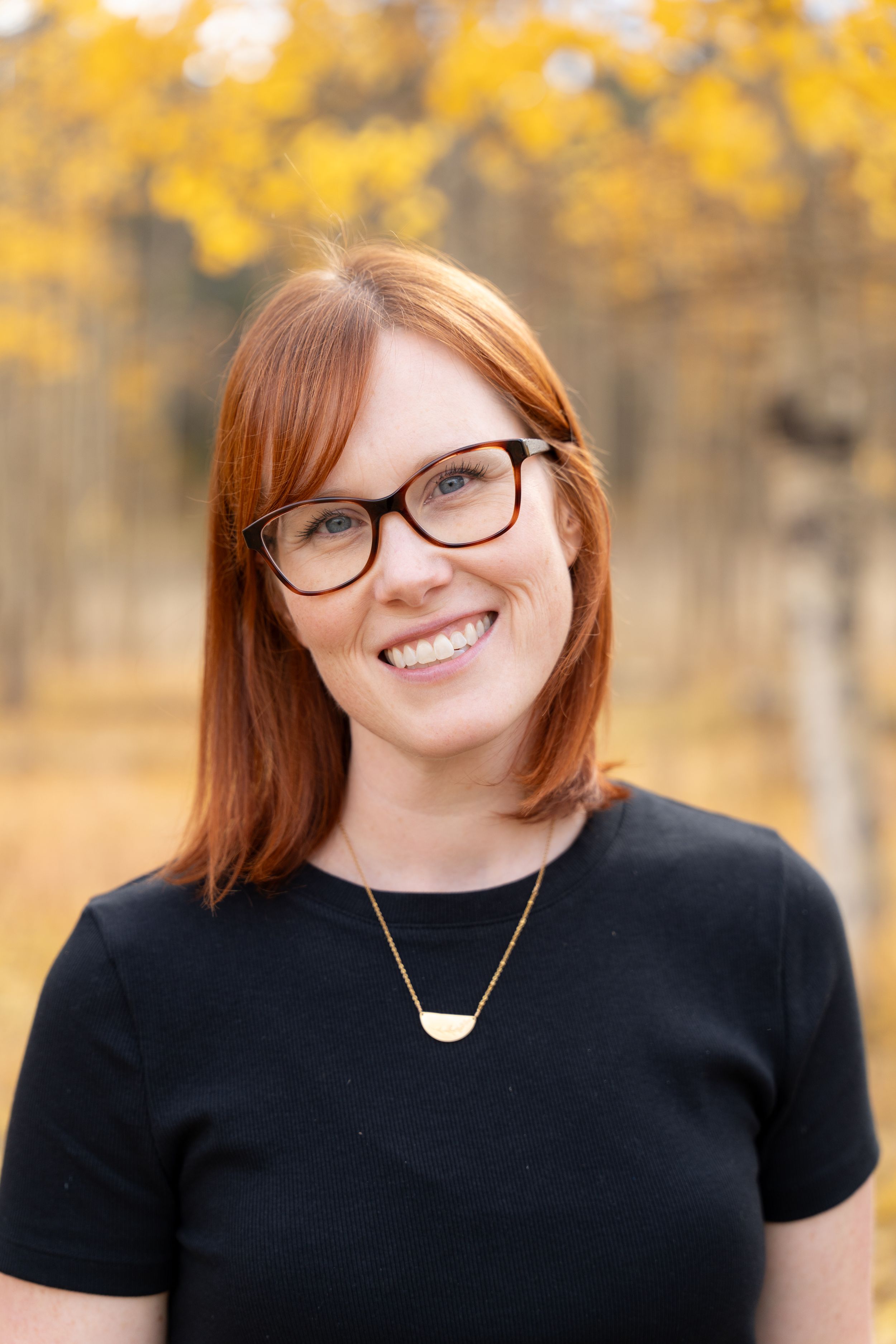 Professional portrait of woman in black shirt and glasses against golden fall foliage background.