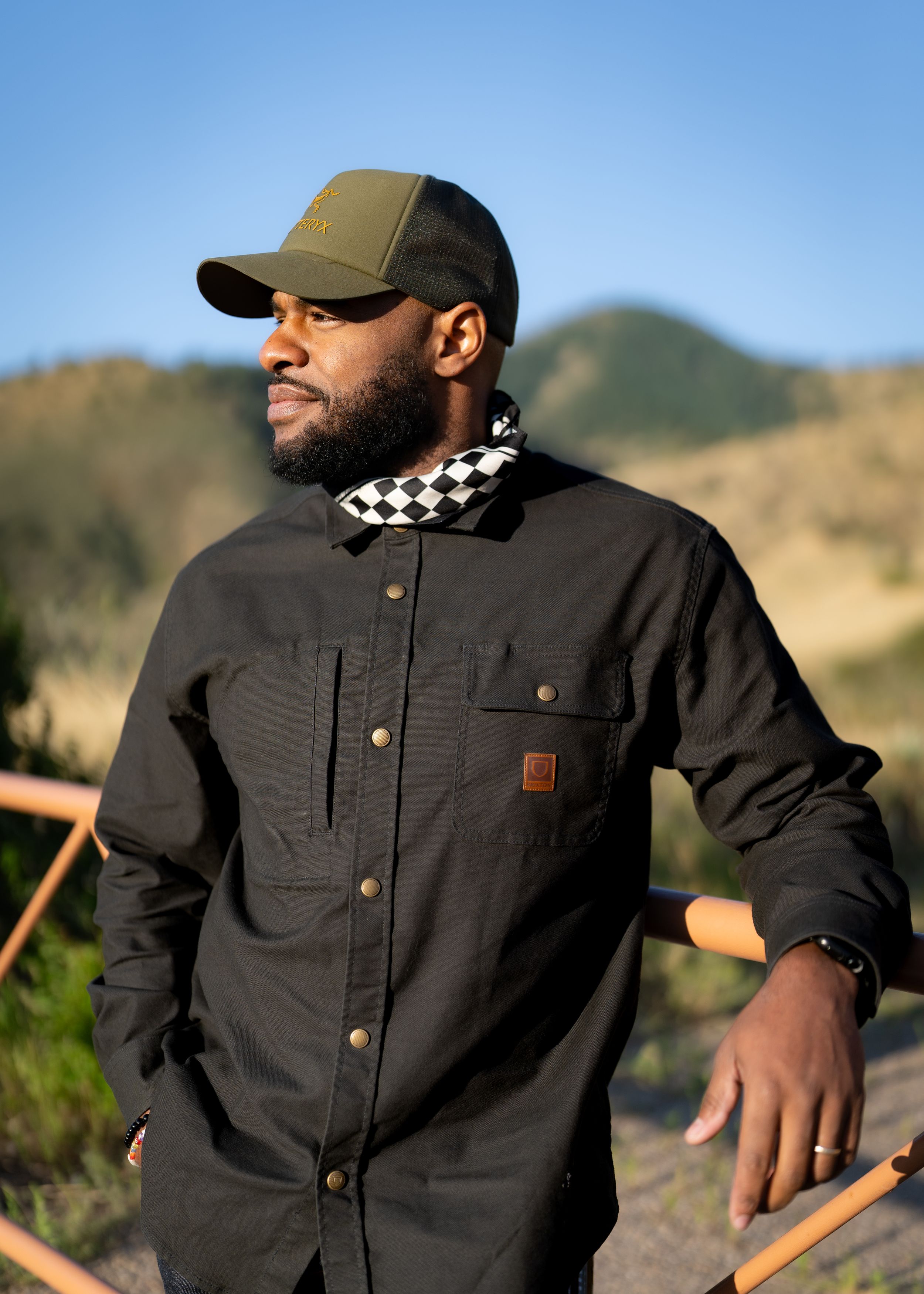 Man in motorcycle jacket with checkered bandana enjoys scenic mountain view at golden hour.