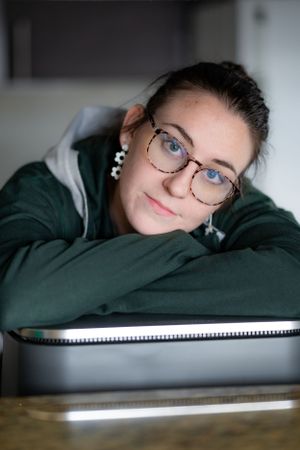 A young woman wearing glasses and a green hoodie rests their head on their folded arms at a countertop.