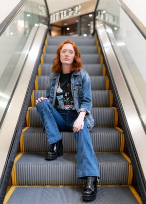Young woman with red hair in denim jacket and jeans poses confidently on mall escalator.