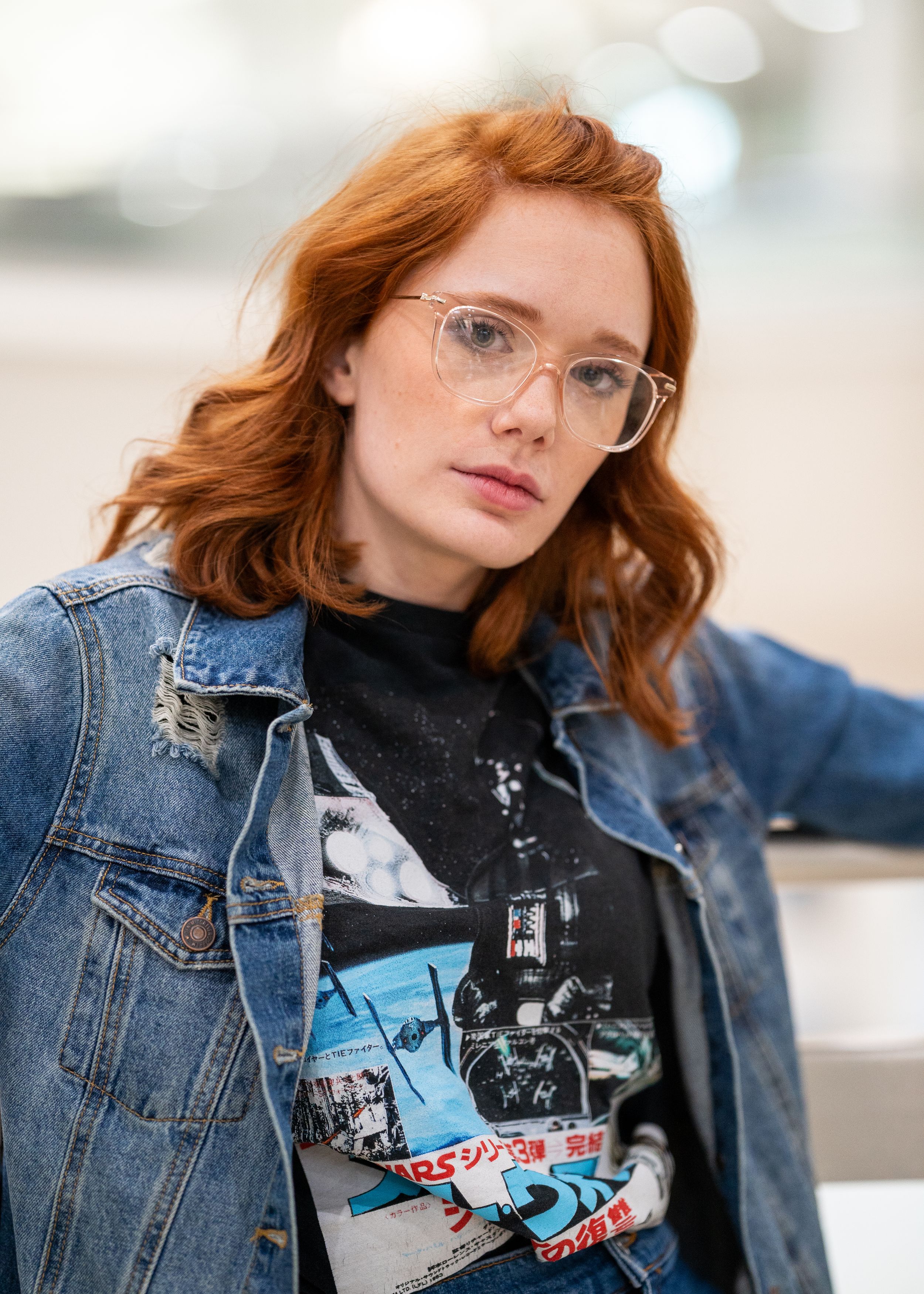 Artistic portrait of young woman with red hair wearing glasses, denim jacket, and graphic t-shirt against bright blurry background.
