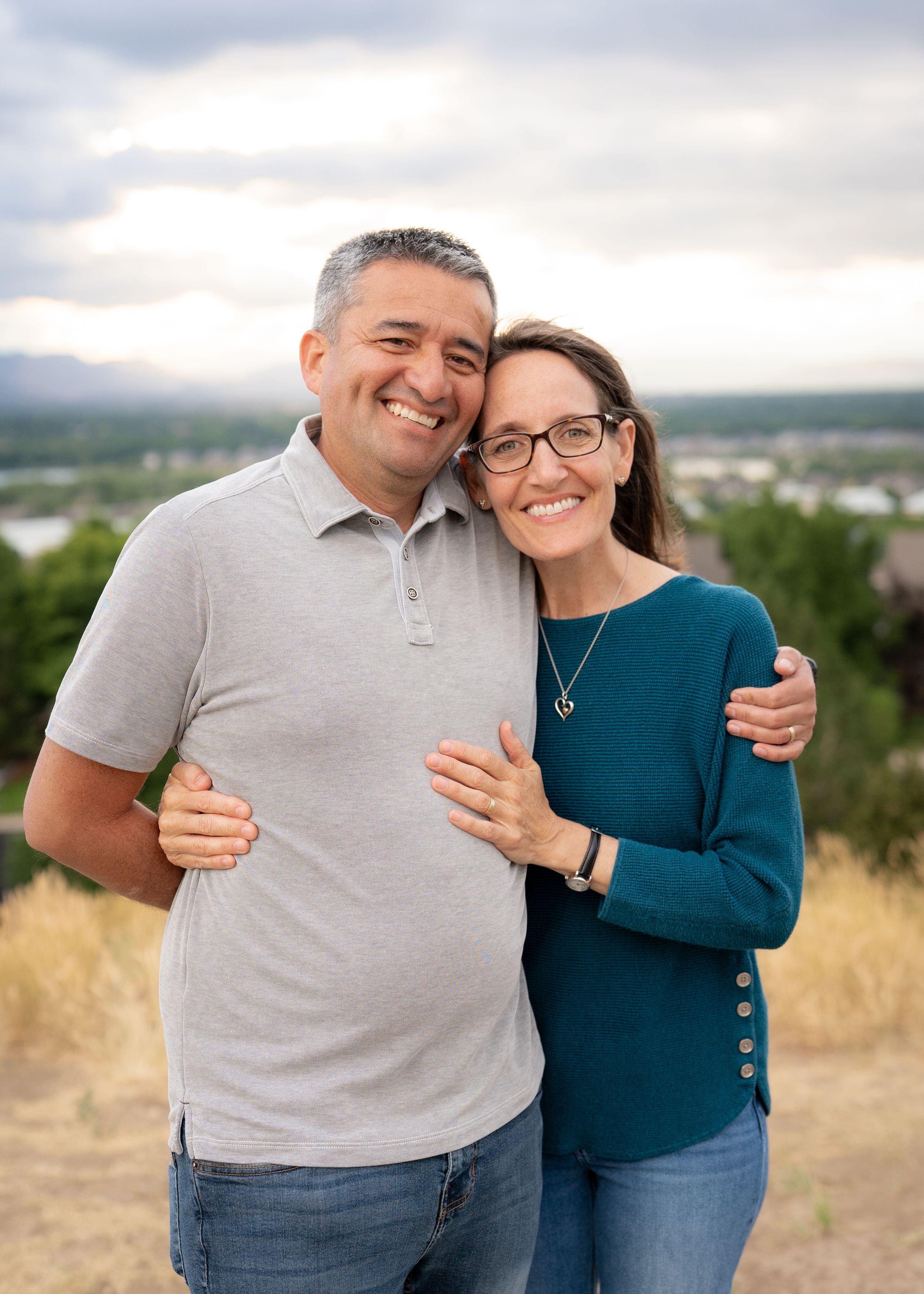 Married couple embraces while smiling at the camera on a hilltop with a scenic view behind them.