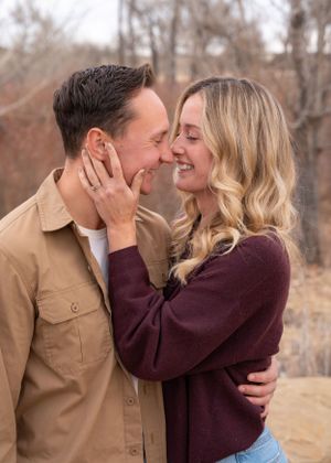 A romantic couple shares a tender moment outdoors in autumn, with the woman in burgundy and man in tan embracing closely.