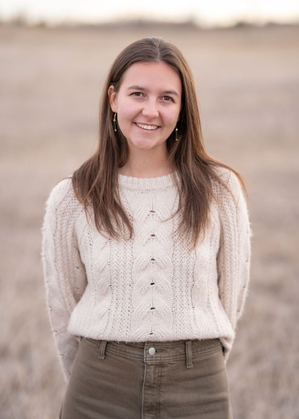 A woman with flowing brown hair poses outdoors wearing a cozy cream-colored cable knit sweater and brown pants.