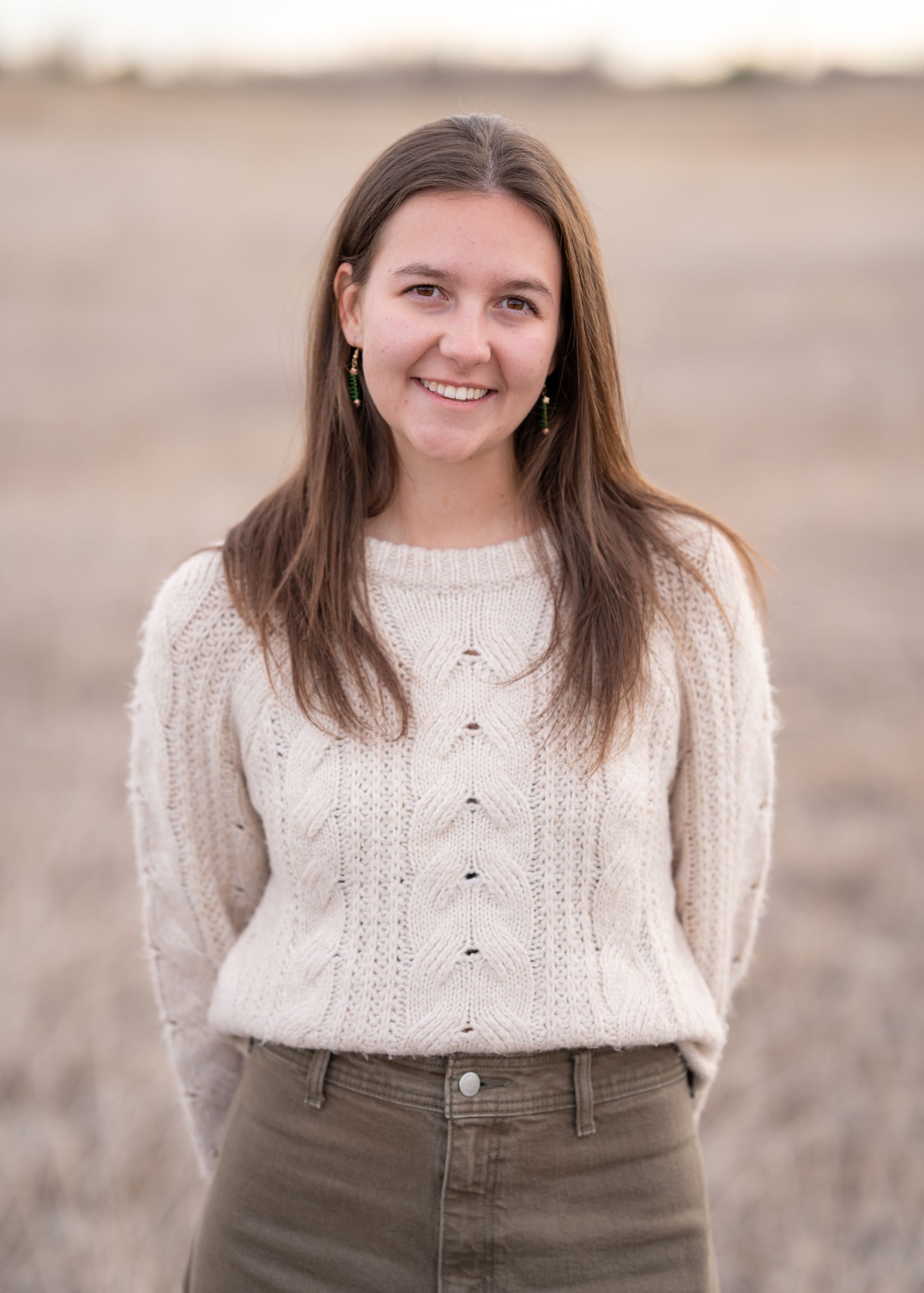 A woman with flowing brown hair poses outdoors wearing a cozy cream-colored cable knit sweater and brown pants.