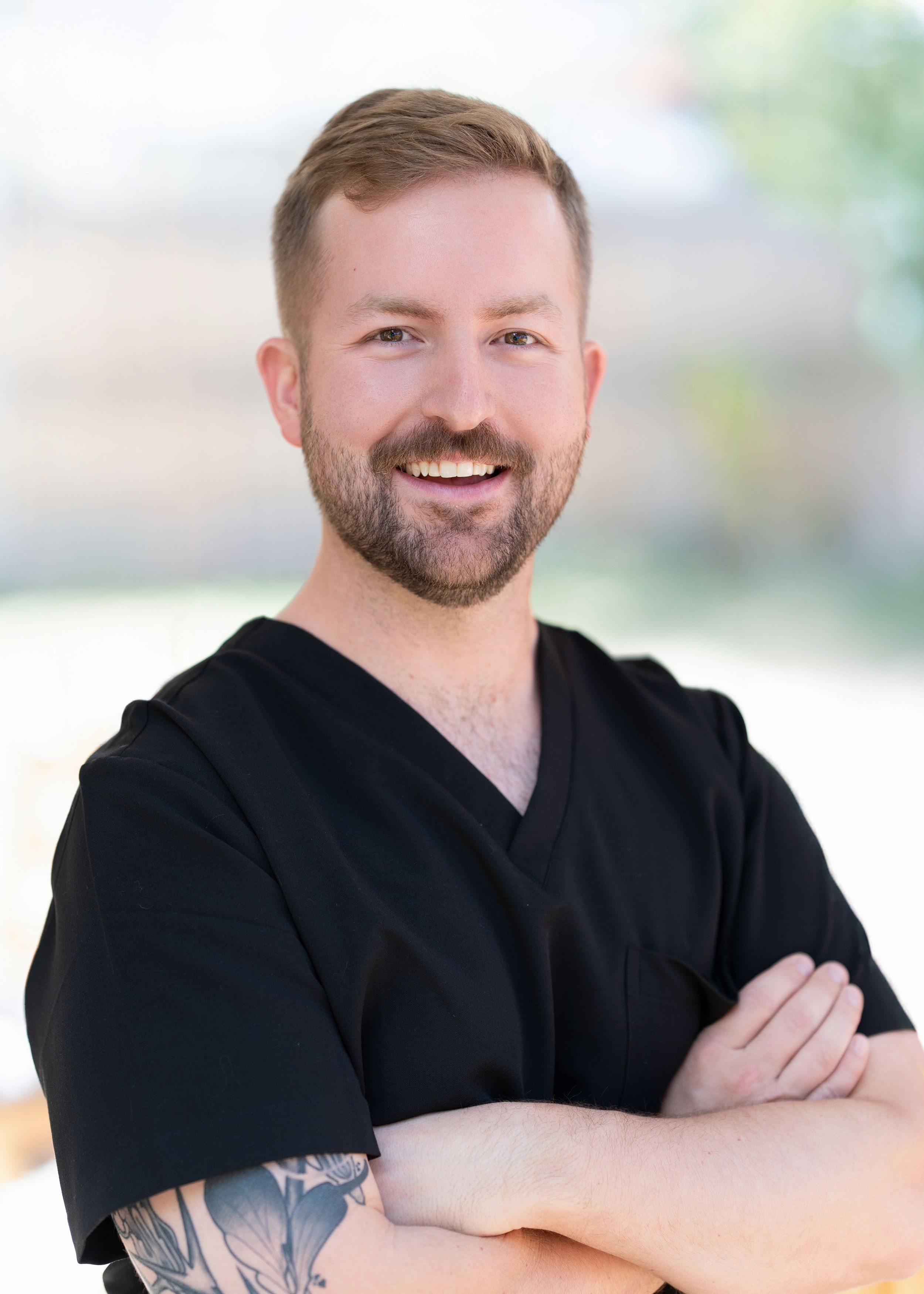 Professional portrait of healthcare professional in black scrubs with tattoo and confident smile.