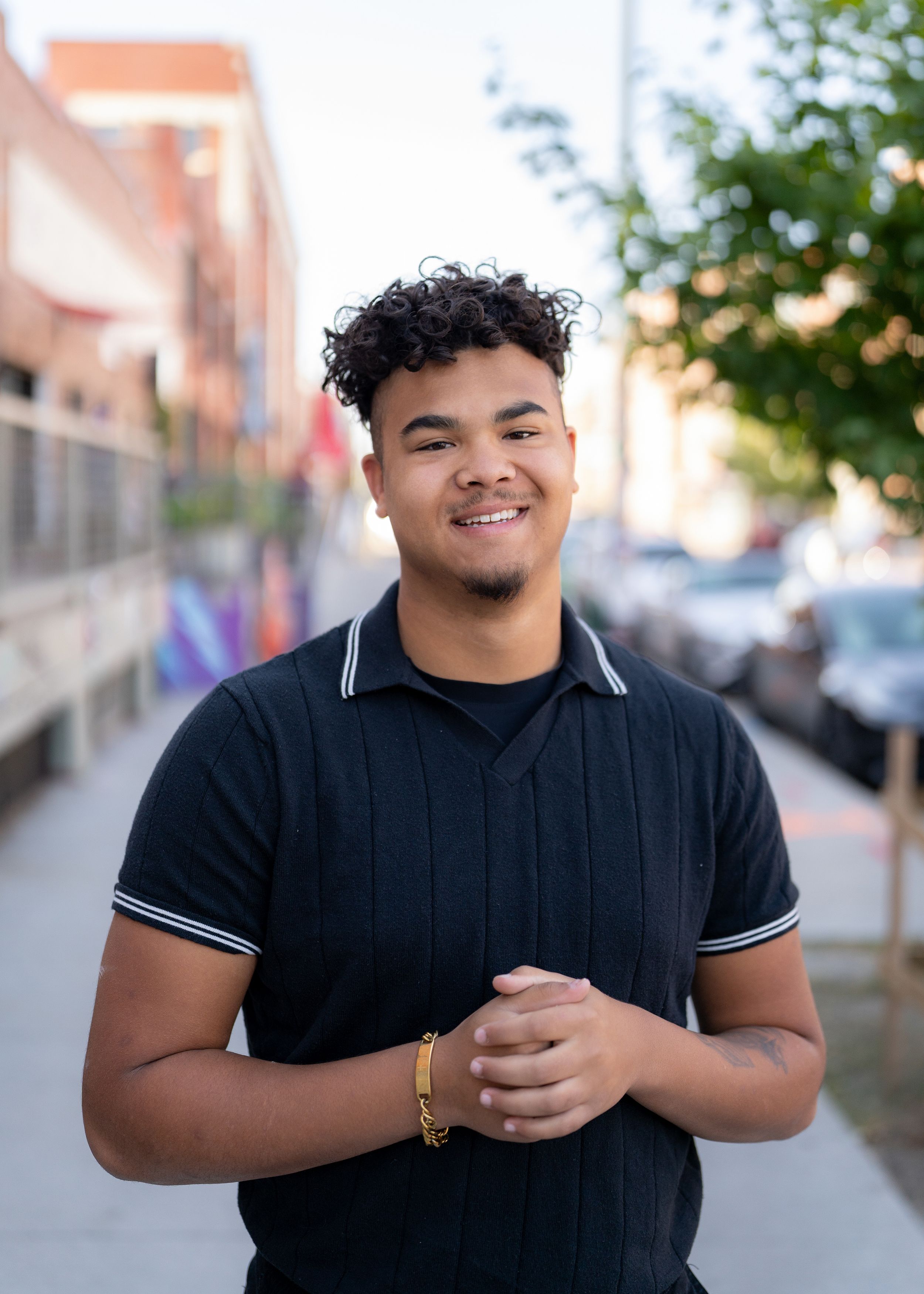 Professional portrait of high school senior in black polo shirt smiling warmly while standing on urban sidewalk.